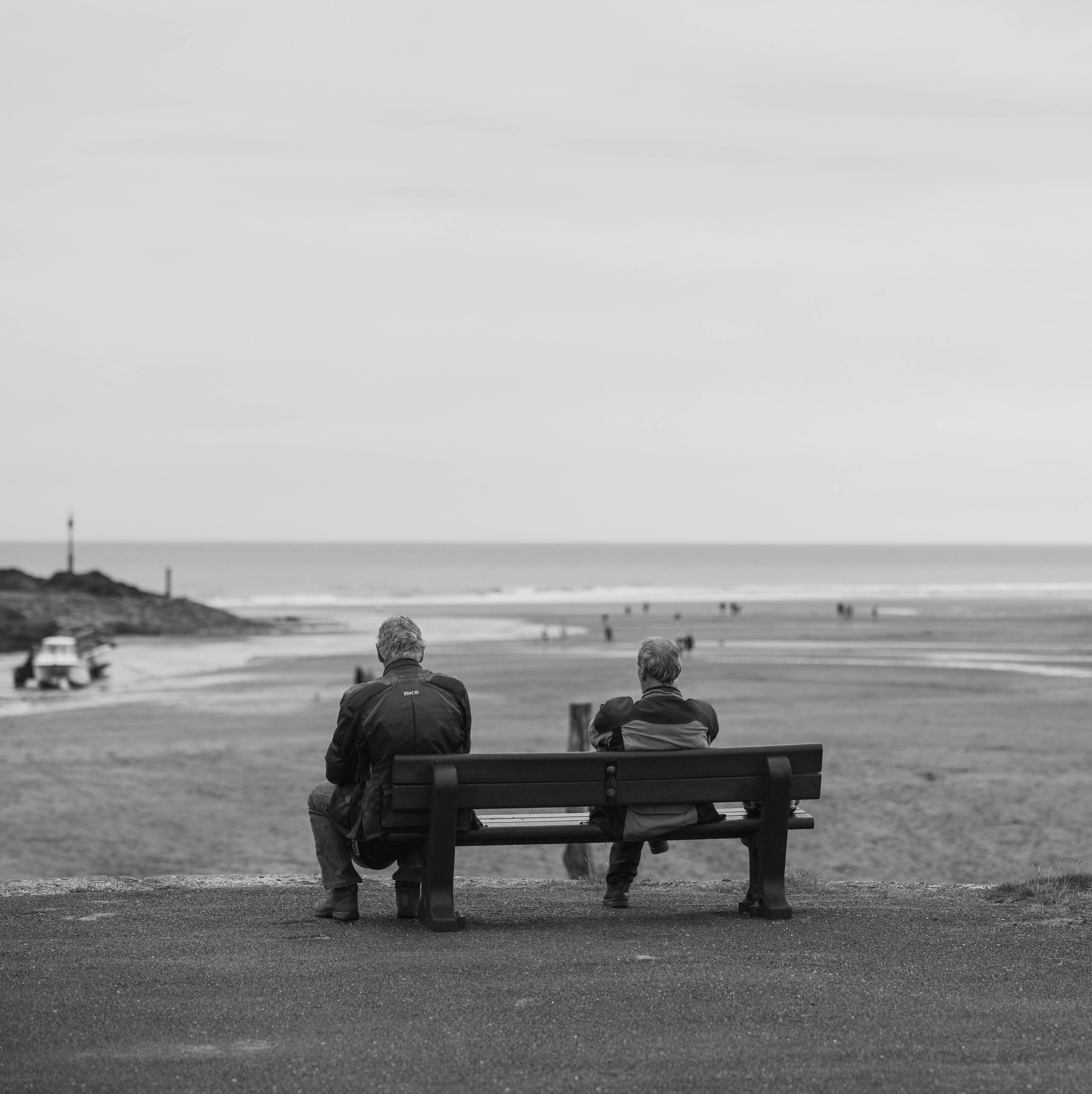 Two people sitting on a bench, quietly facing a broad beach and calm sea.